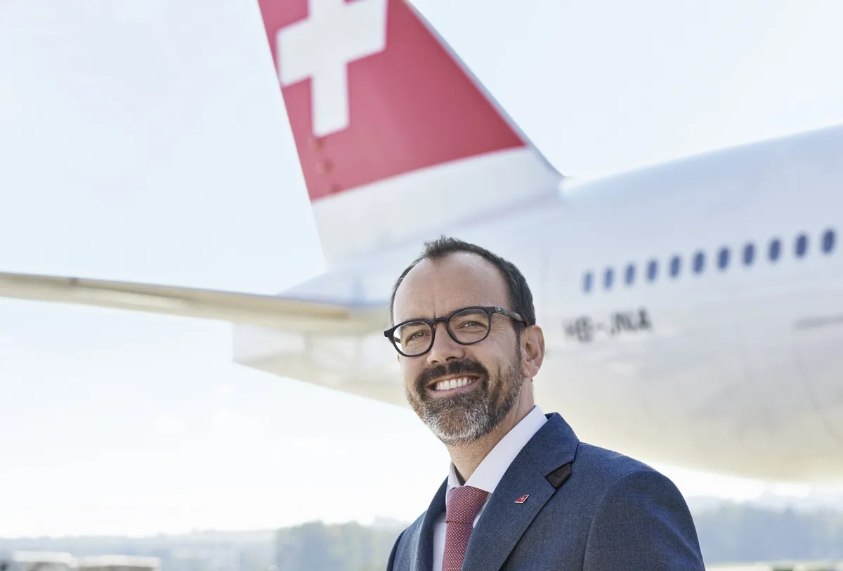 Ein Mann mit Brille und Bart lächelt in die Kamera. Im Hintergrund die Heckflosse eines Swiss-Flugzeugs.